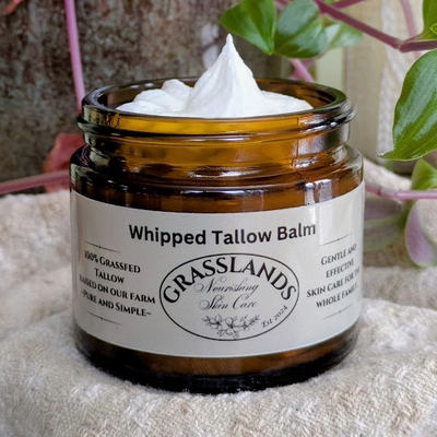 Jar of Grasslands Whipped Tallow Balm with a white label on a textured surface with plants in the background.