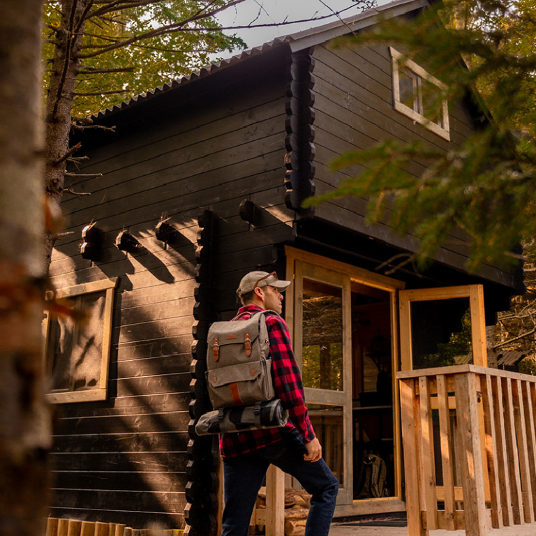 Person with a backpack standing on the steps of a wooden cabin in a forest setting