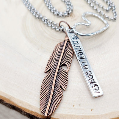 a necklace with a feather, a heart and "Beautifully Broken" stamped on it