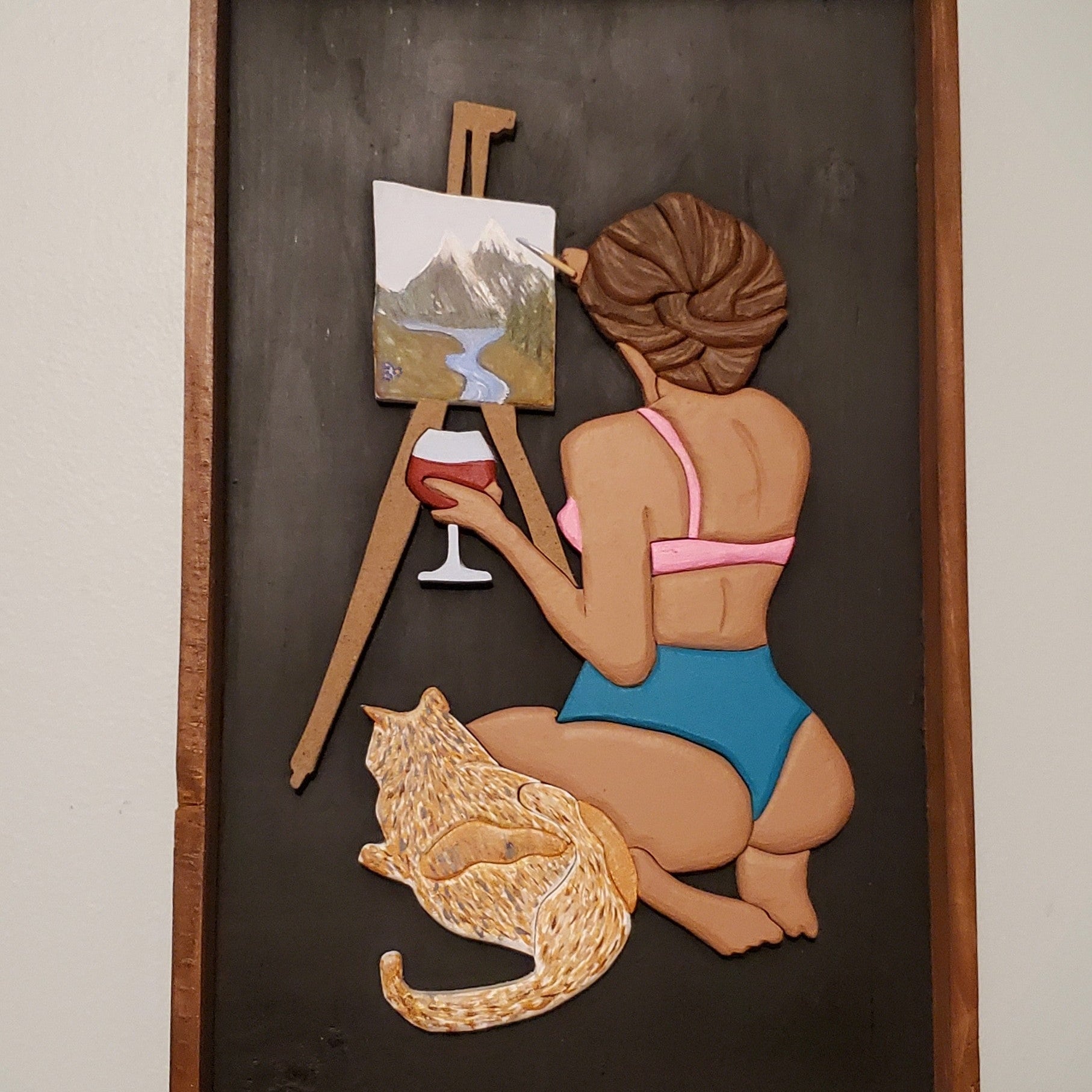 Decorative wall art of a woman in a bikini painting a landscape with a cat at her feet.
